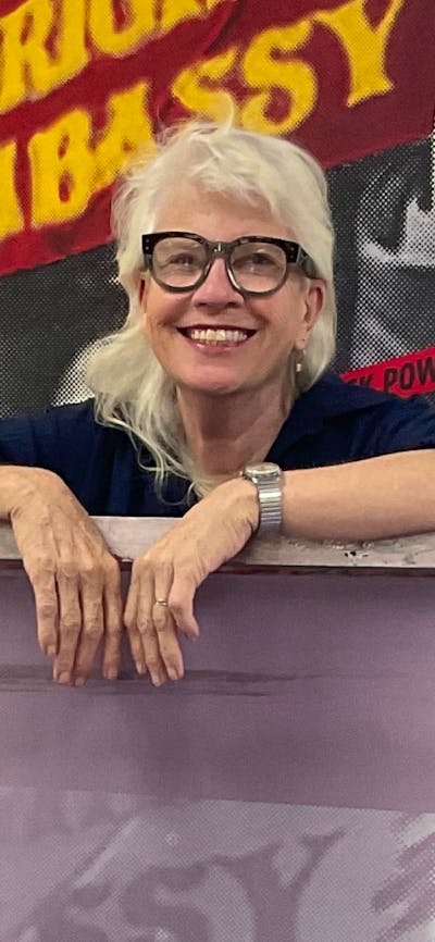 A woman with blonde hair and glasses leaning on a purple artwork piece.