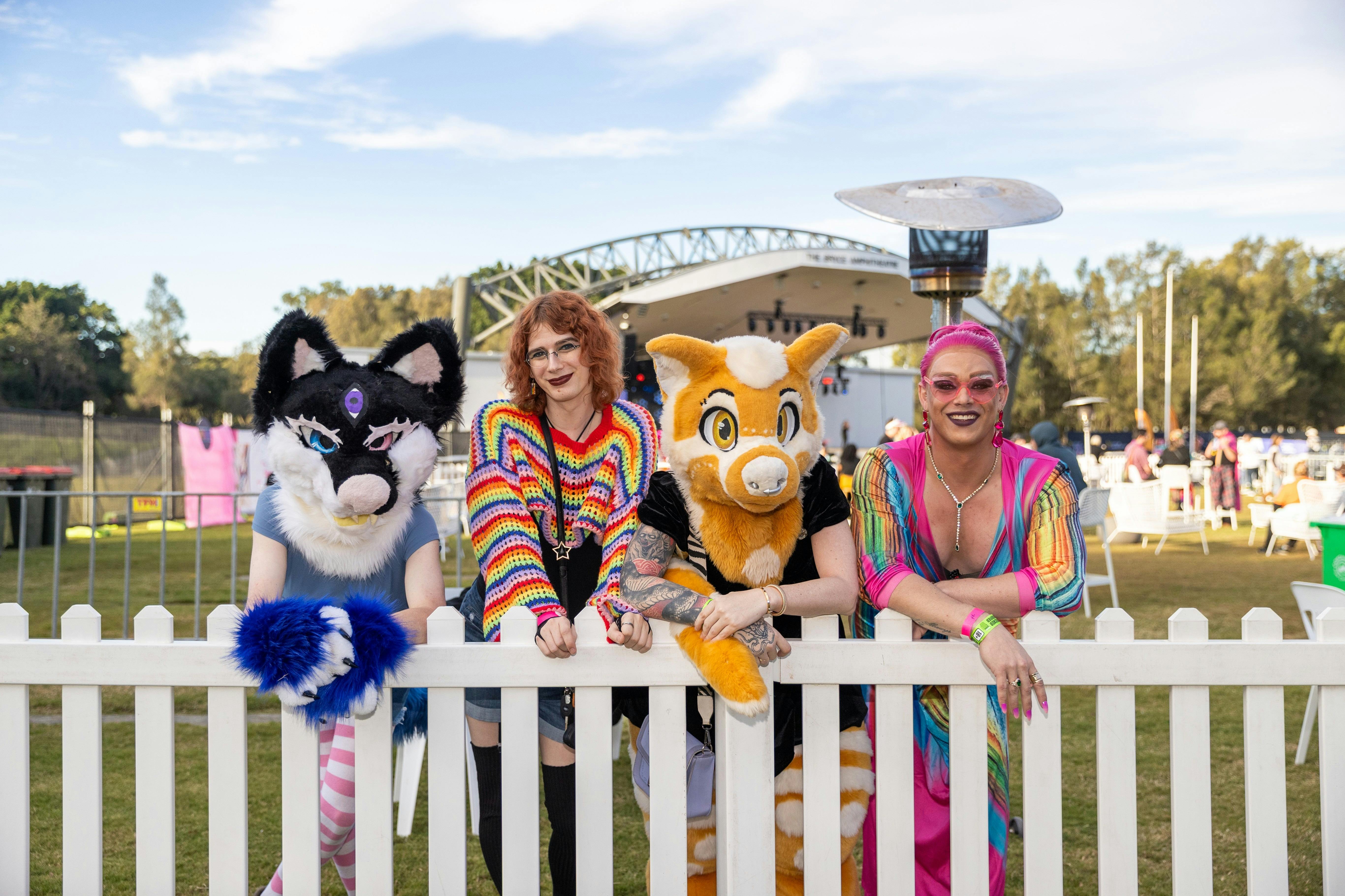 Four attendees pose behind a white picket fence at Moreton Bay PrideFest. Two are wearing colourful