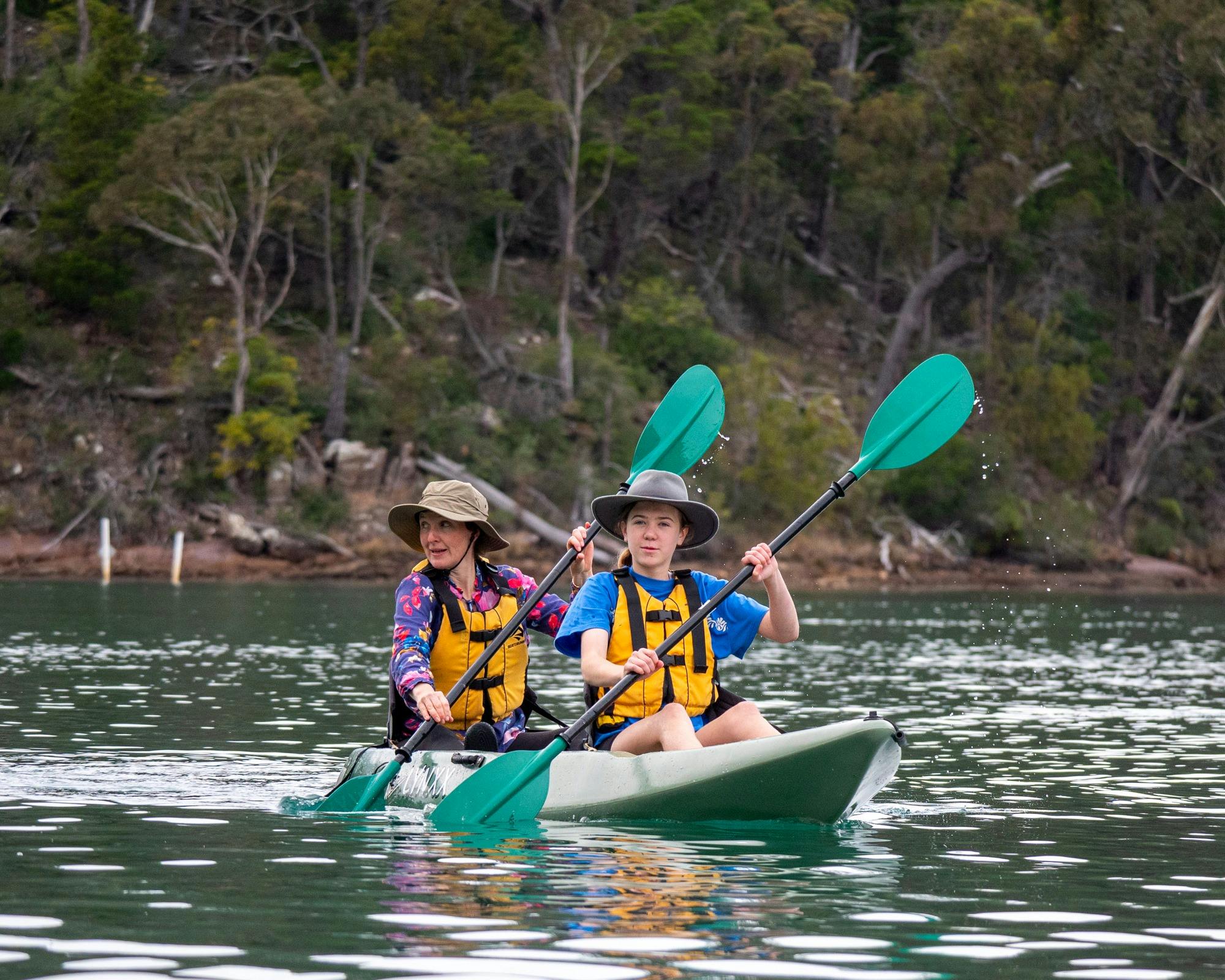 A mother and daughter in a green kayak, wearing hats and PFDs, smiling as they paddle in sync on the