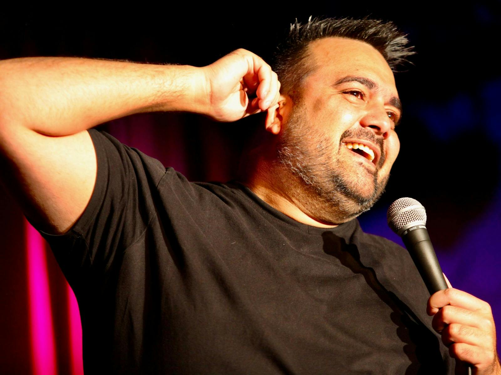 Aboriginal man in a black t-shirt holds a microphone in one hand and has his other arm  raised