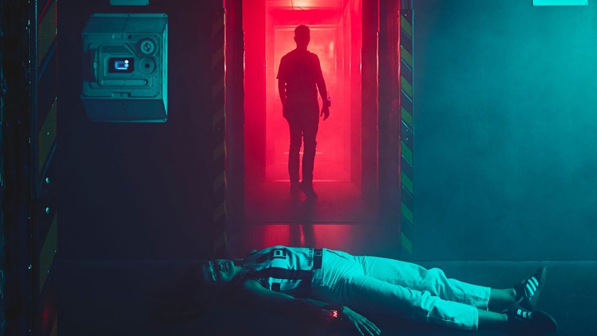 A player lies on the floor of a red-lit corridor as a silhouetted figure walks away.