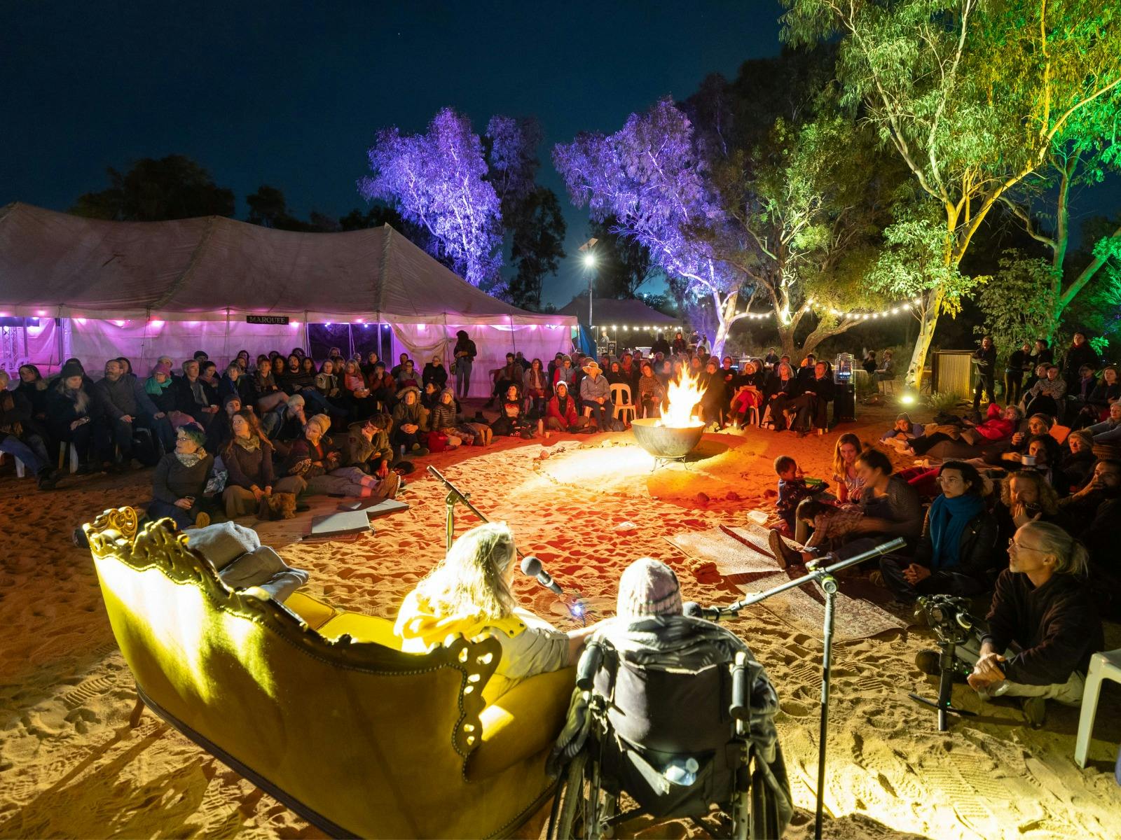 An audience gathers at night around a fire, listening to speakers under string lights and trees.
