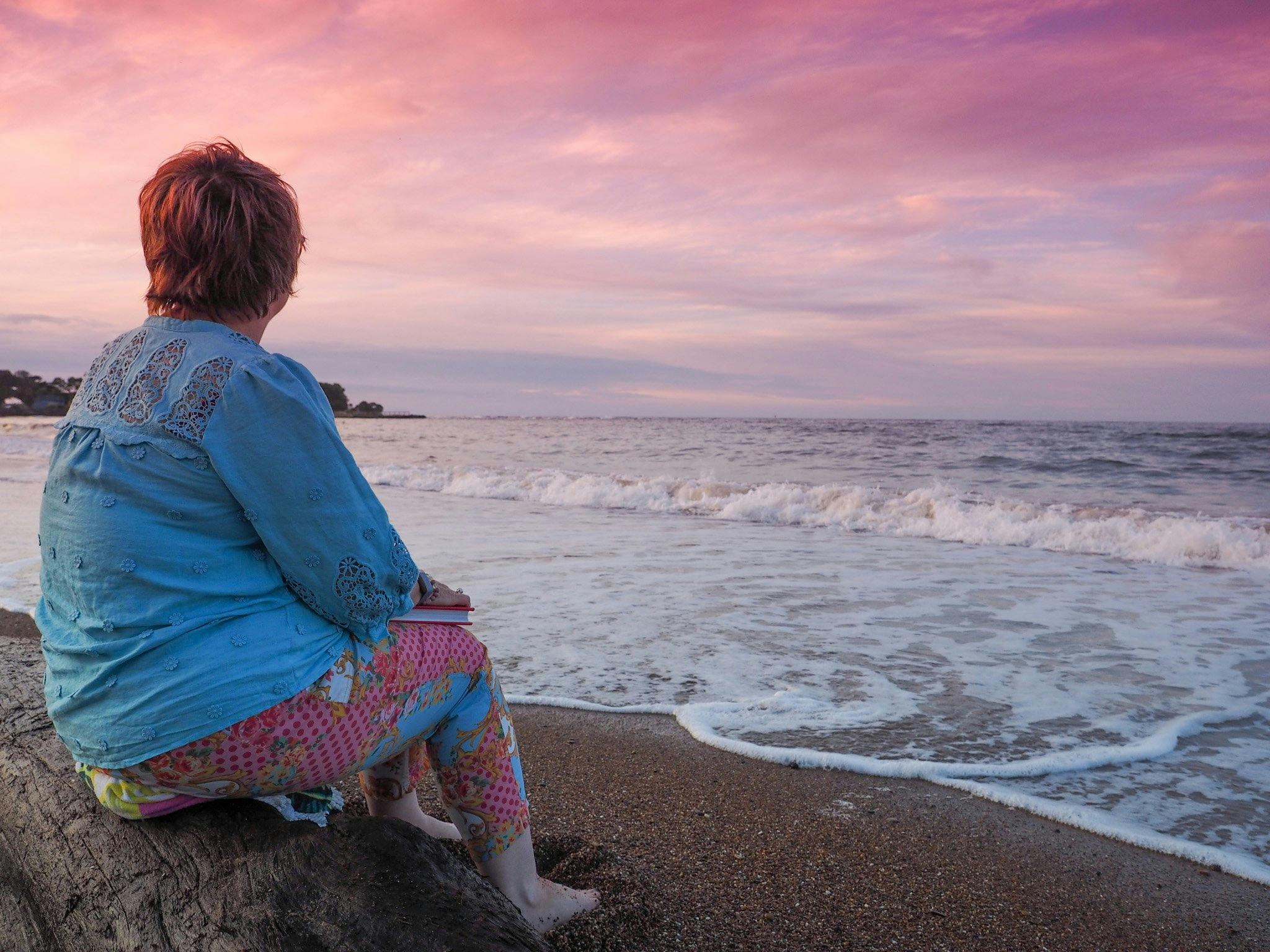 Woman sitting on log on beach, looking out across the horizon of where the ocean meets the sunset.