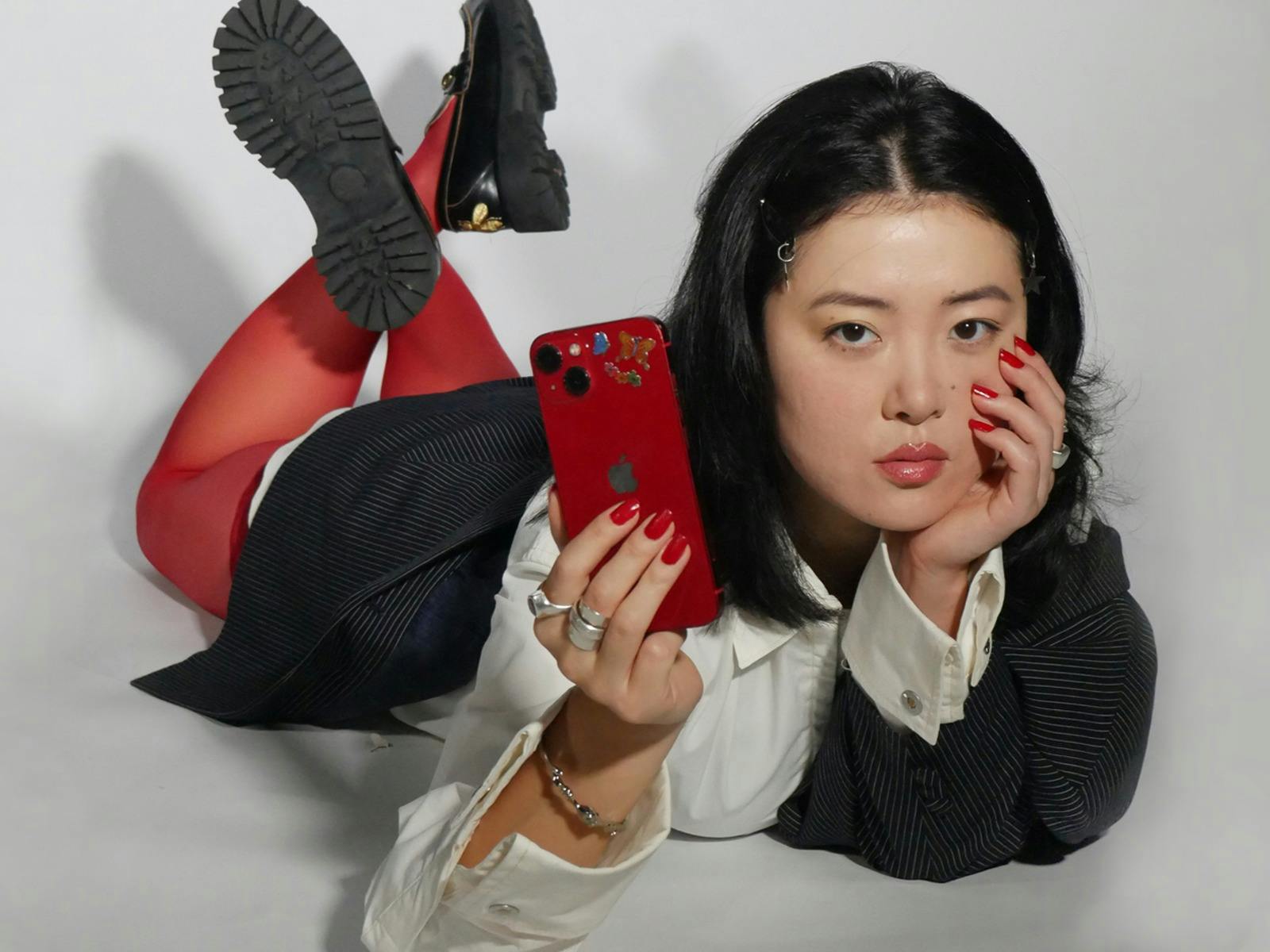a young fashionable woman lies, legs crossed behind her, with her phone pointing out at the viewer