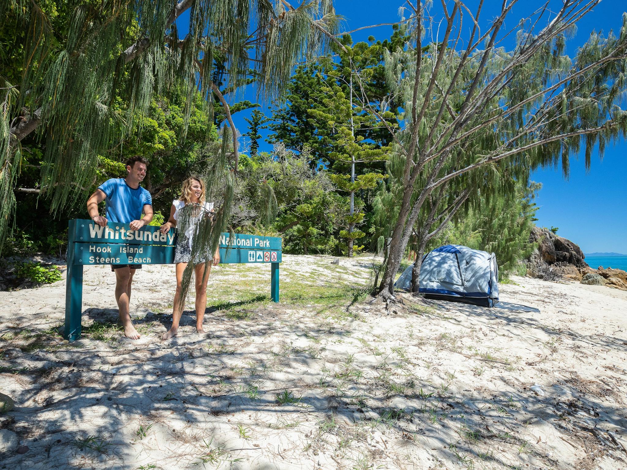 Smiling couple standing at a Hook Island, Steen Beach sign with a camping tent nearby Smiling couple standing at a Hook Island, Steen Beach sign with a camping tent nearby