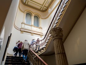 5 adults walk up a large ornate staircase with high ceilings in the Hellenic Museum foyer