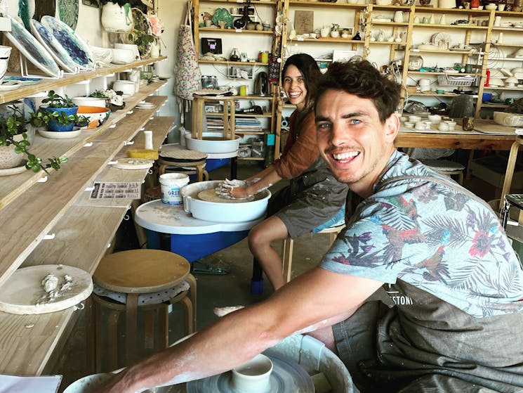 photo shows two people looking at the camera and smiling while they sit at a pottery wheel