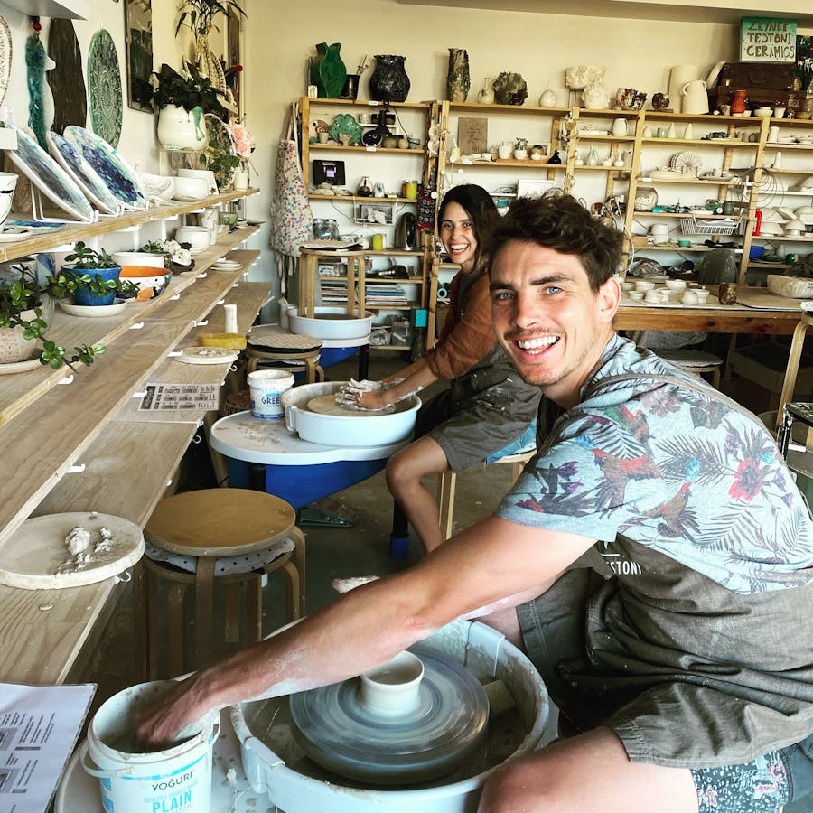 photo shows two people looking at the camera and smiling while they sit at a pottery wheel