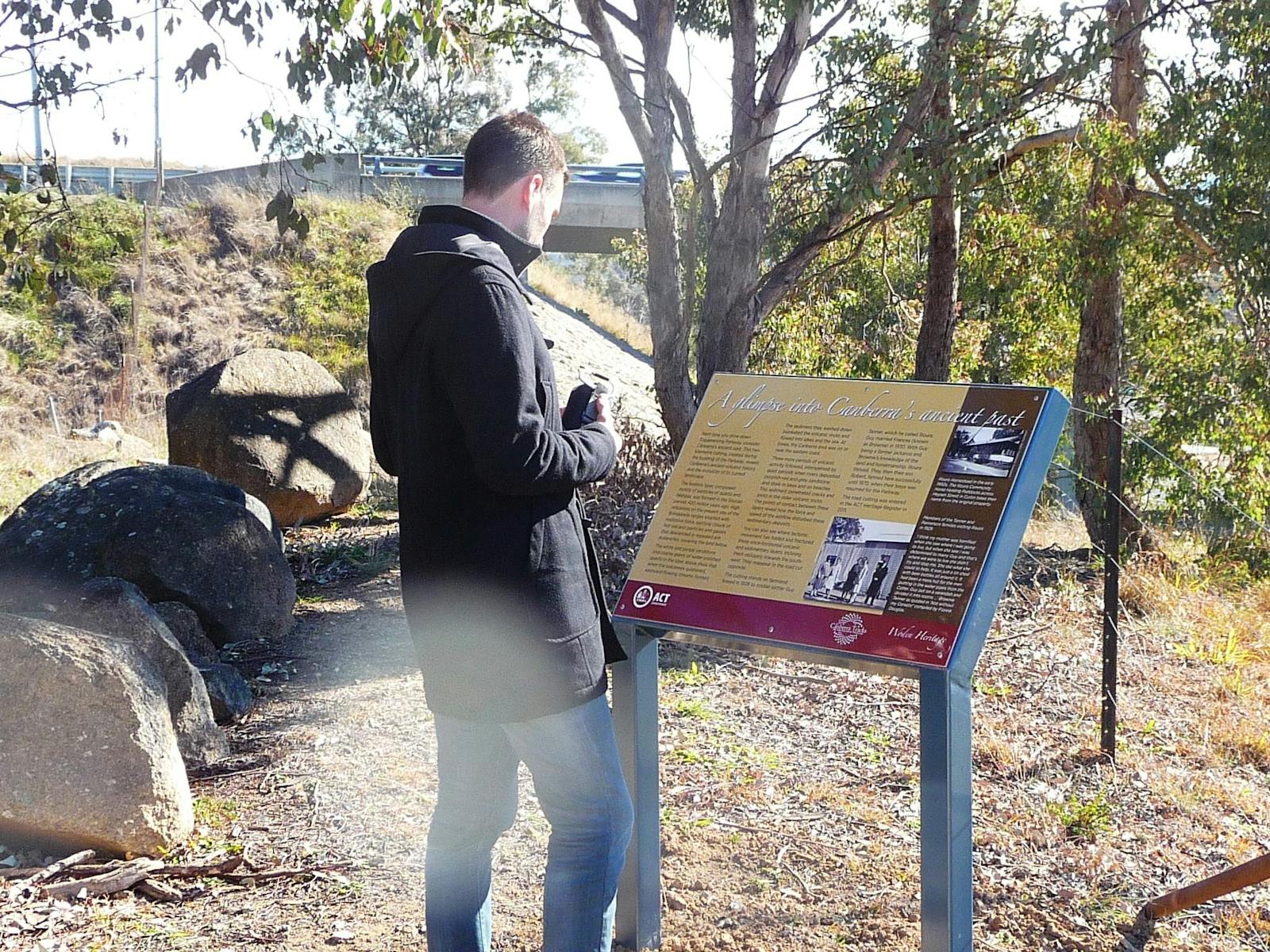 Man reading a sign board in treed area with road bridge in background