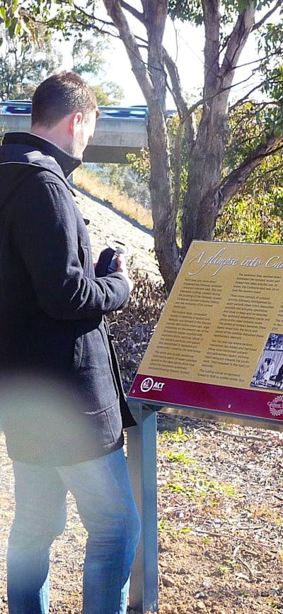 Man reading a sign board in treed area with road bridge in background