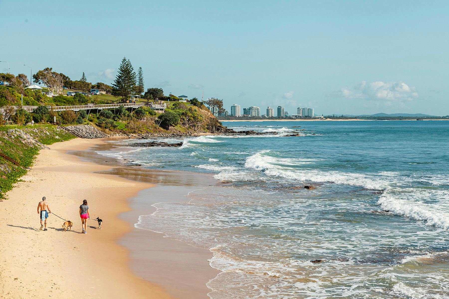A couple walk their dog along a sandy stretch of beach, facing a headland.