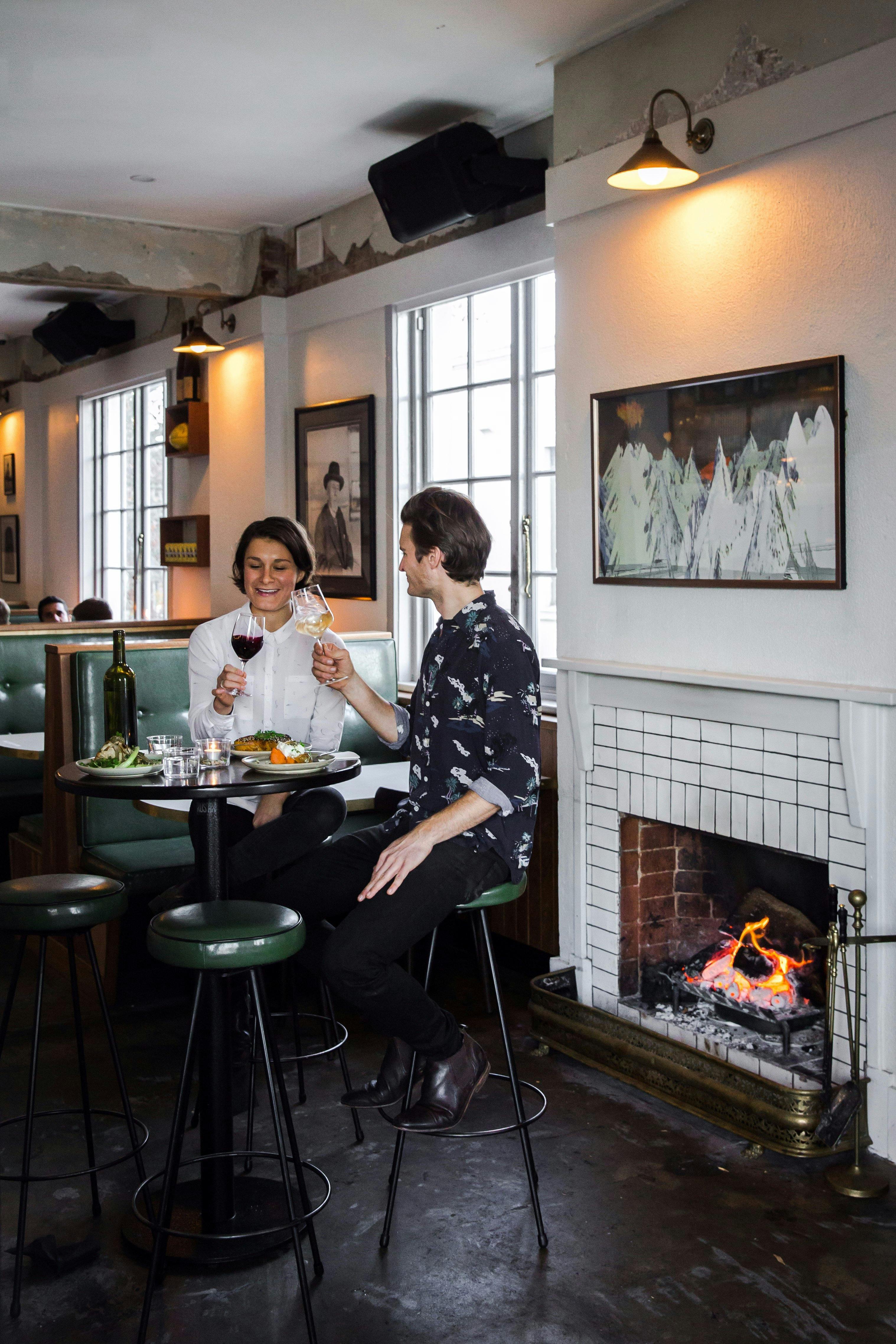 Couple with food and wine on tall stools by a fire place
