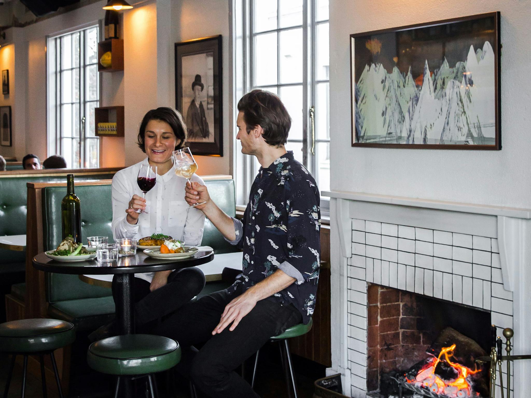 Couple with food and wine on tall stools by a fire place