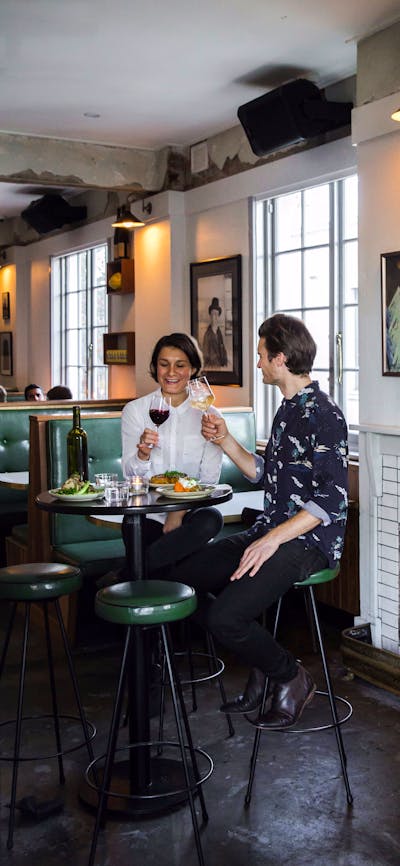 Couple with food and wine on tall stools by a fire place