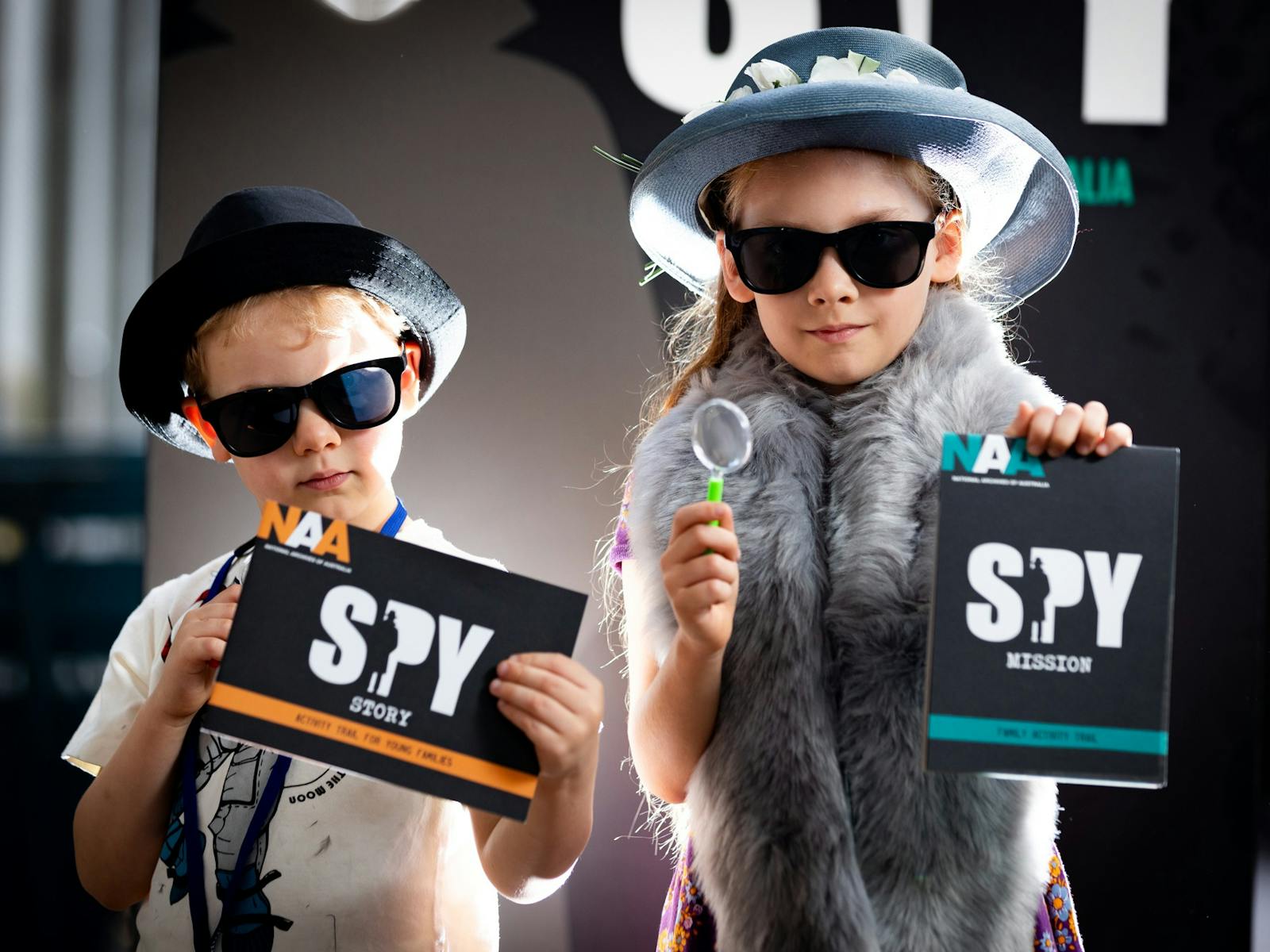 Young boy and girl dressed in spy costumes hold spy-themed activity booklets and magnifying glasses