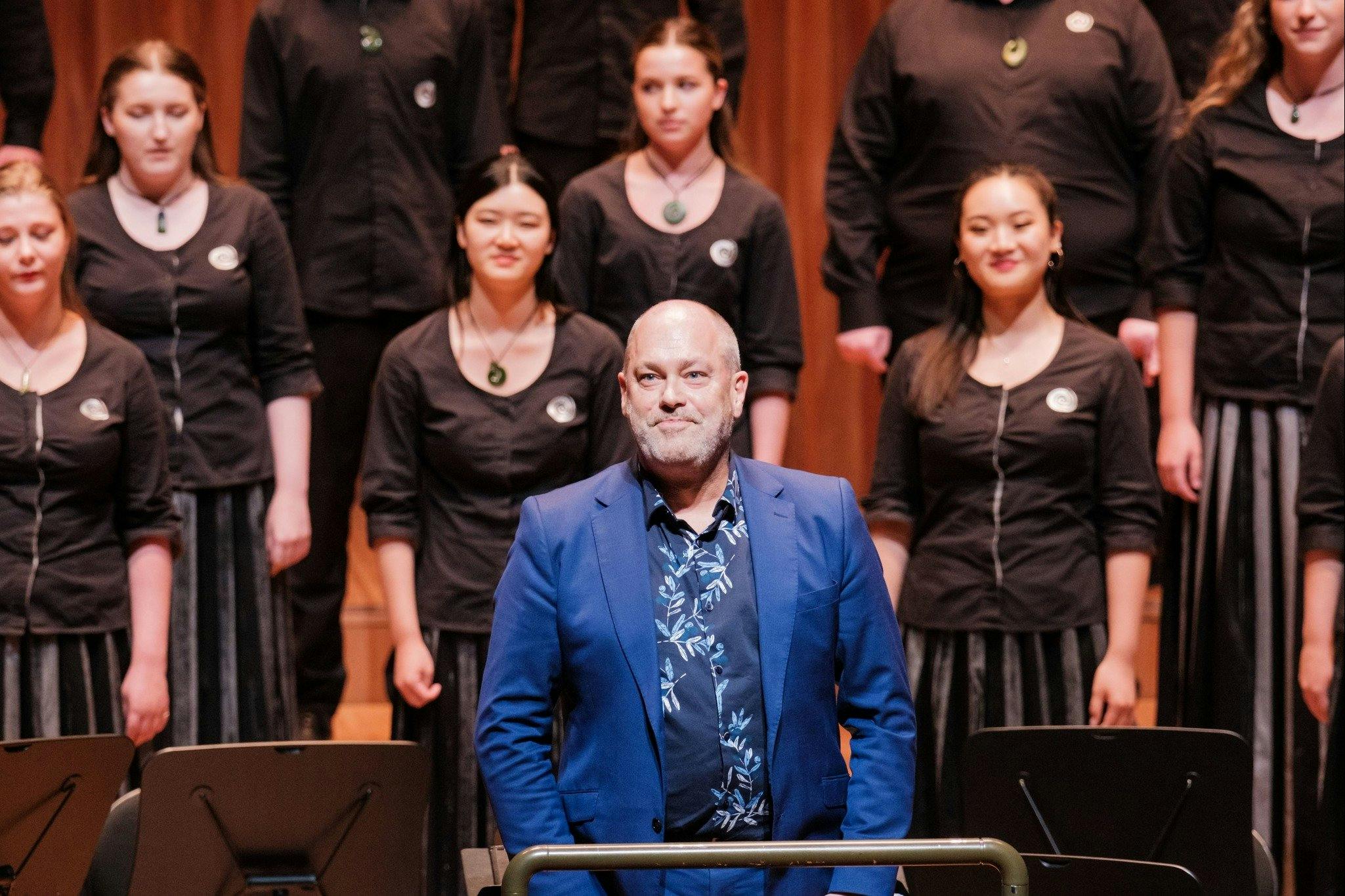 A medium shot of choral conductor David Squire standing in the foreground, wearing a vibrant blue su