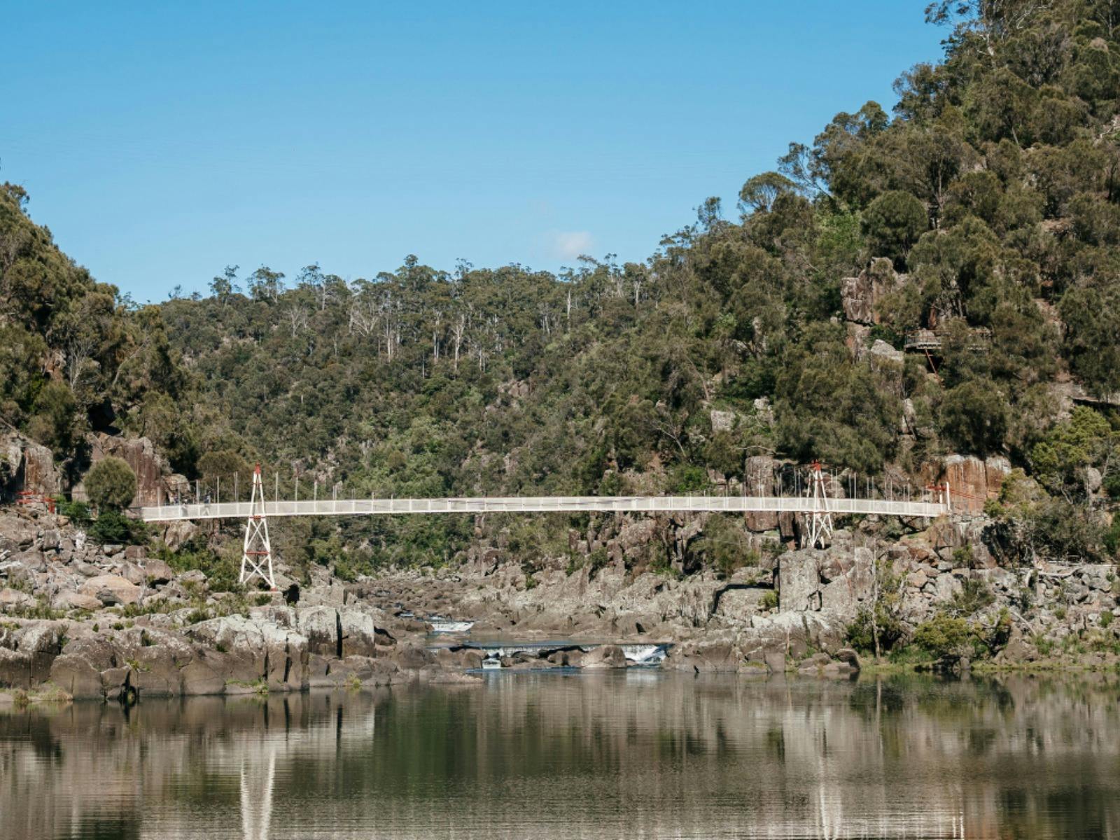A white footbridge over a rocky river gorge, framed by densely wooded hills.