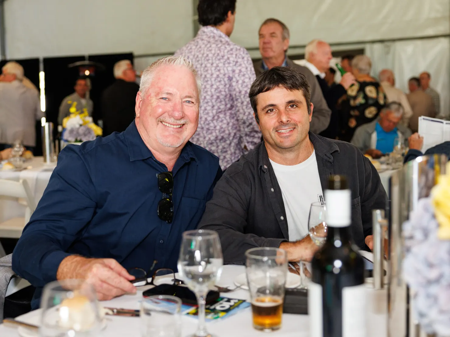 Two men sitting at a table in a marquee smiling