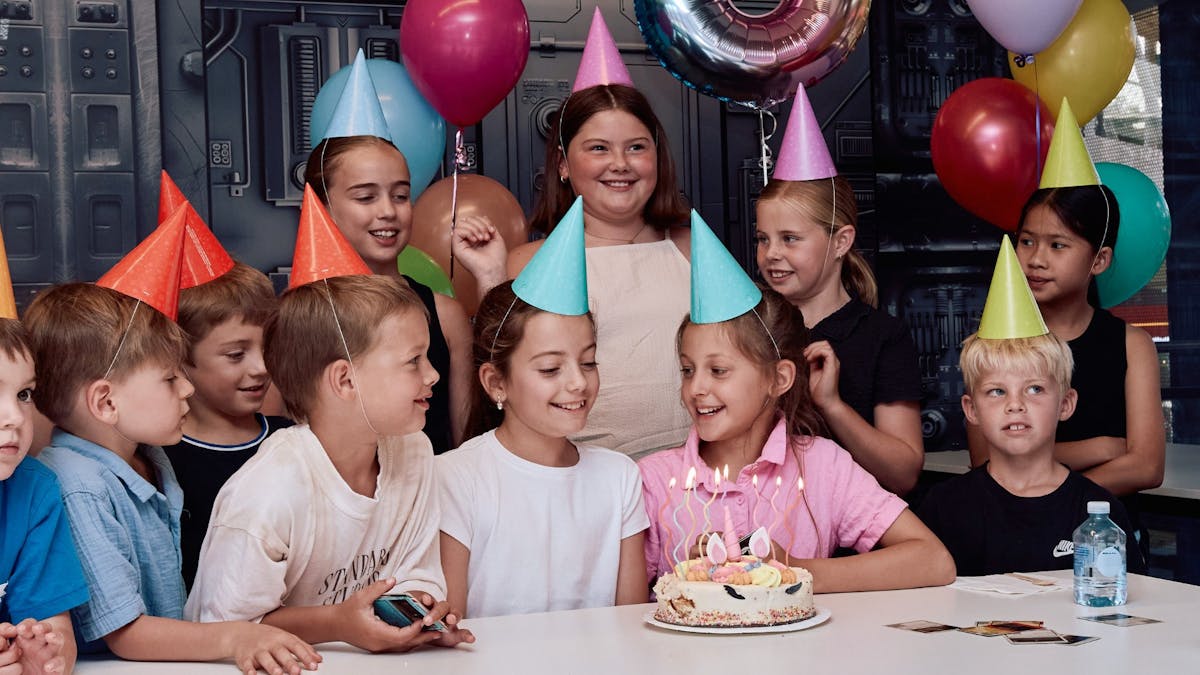 Children in party hats gather around a birthday cake inside a decorated party room. Children in party hats gather around a birthday cake inside a decorated party room.