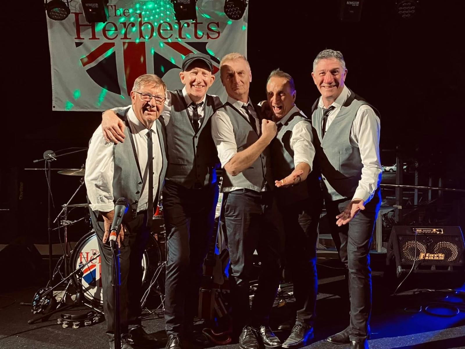 five members of The Herberts standing arm in arm facing the camera and smiling