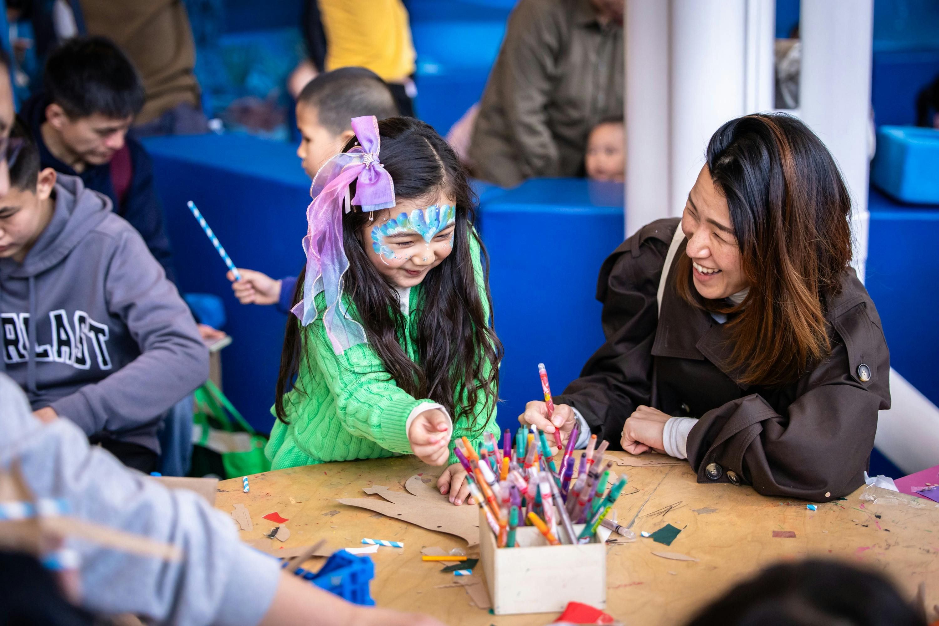 A smiling young girl with face paint and a hair ribbon does crafts at a table with a woman.
