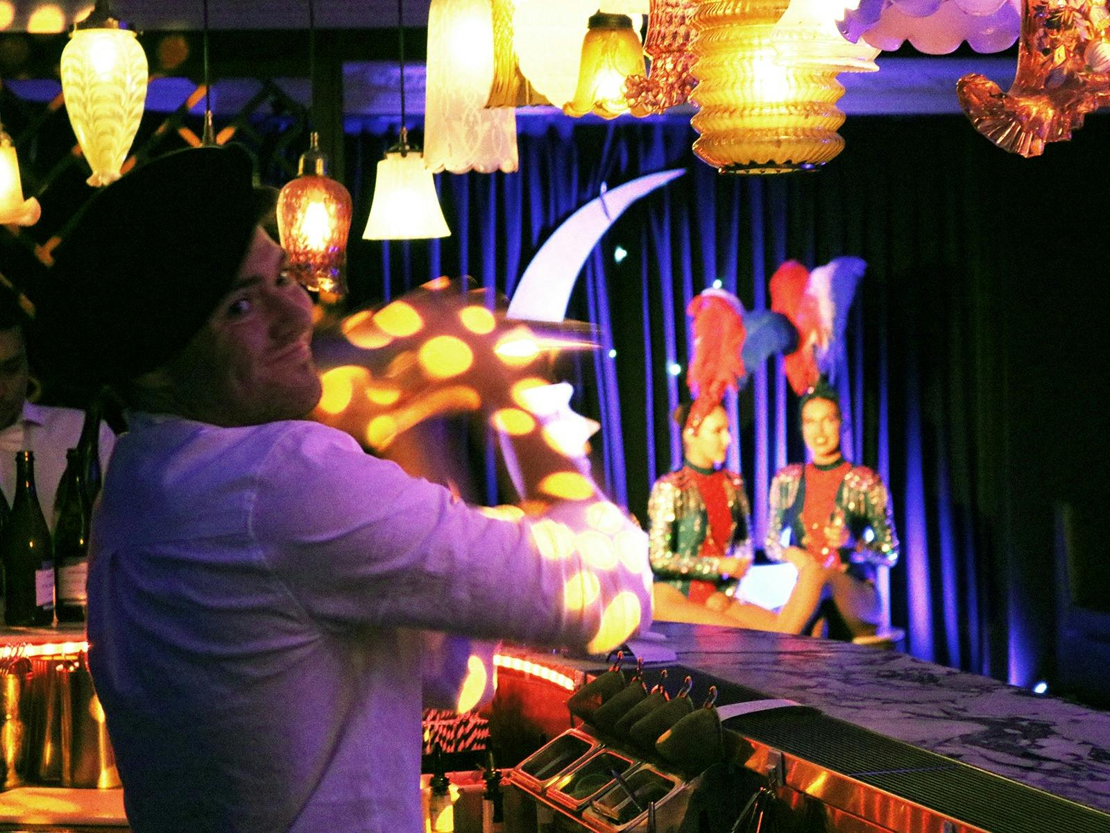A bartender behind bar mixes a cocktail while two showgirls sit in the background in a cabaret bar.