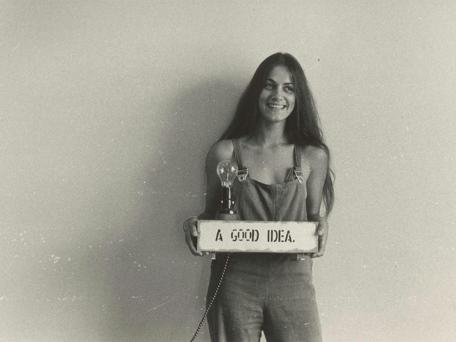 A young woman with long hair holding a sign that says 'A Good Idea' with a lightbulb on top of it.
