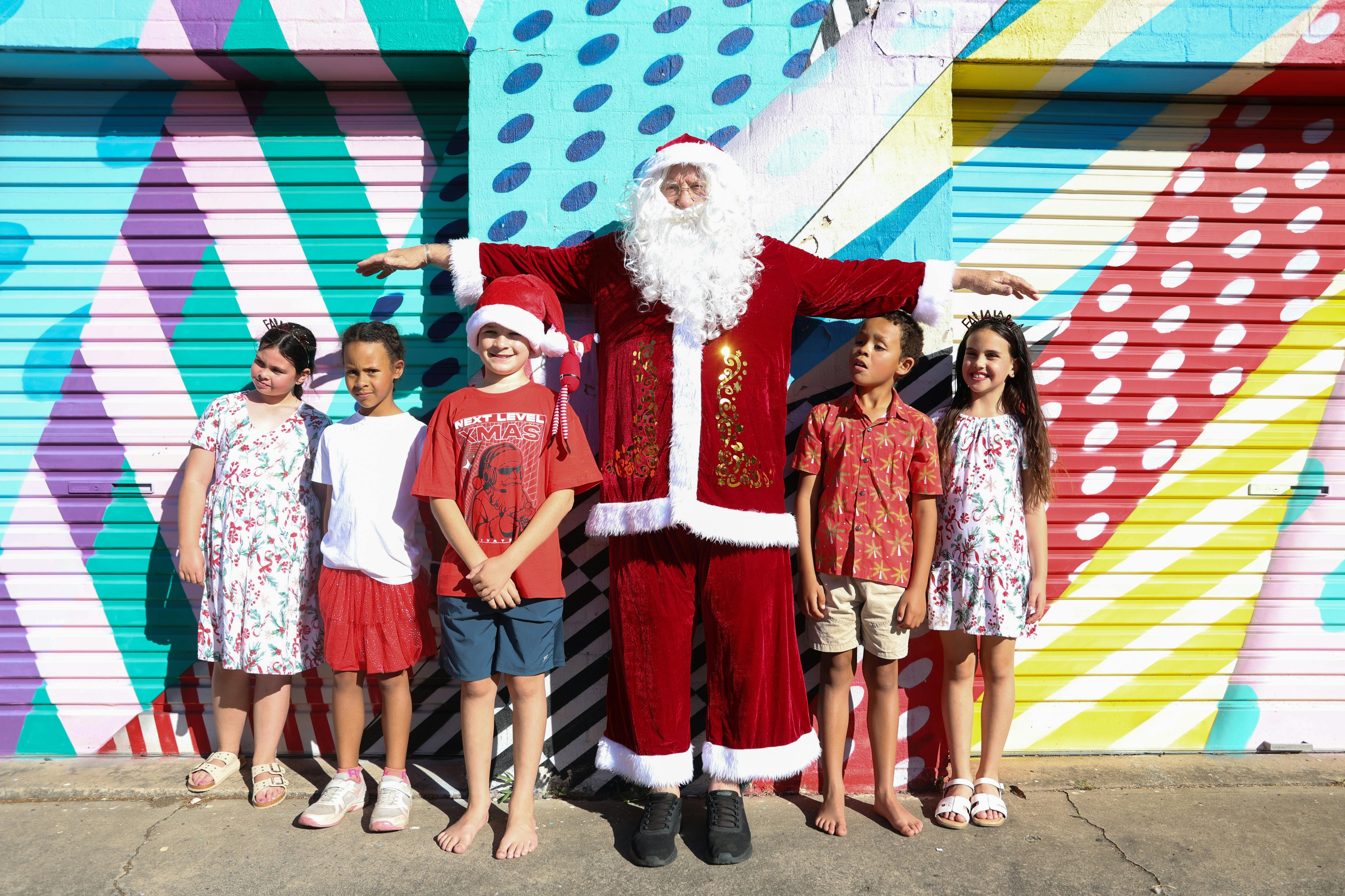 Santa stood in front of a colourful garage holding his arms above the heads of children