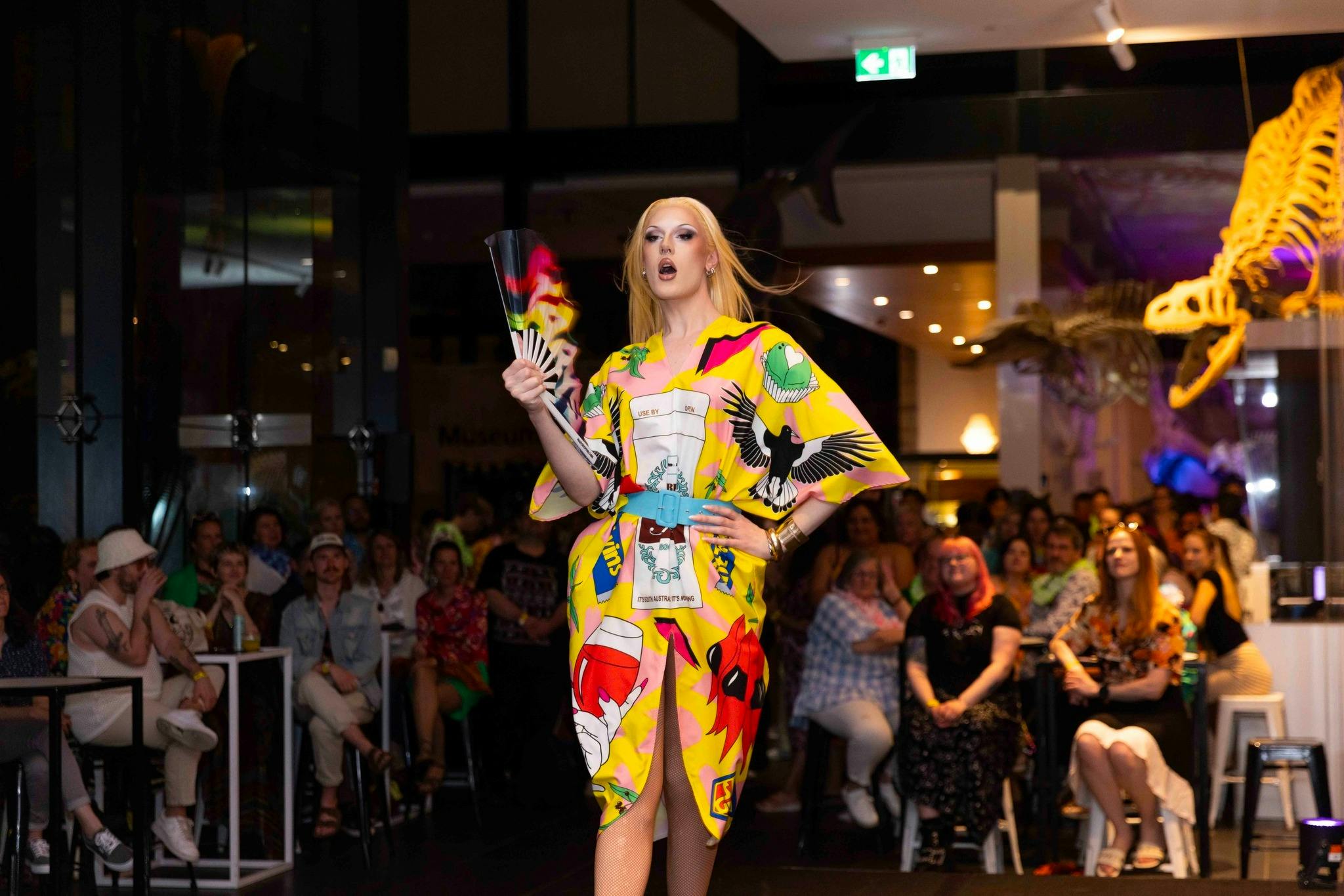A performer in a colorful outfit walks the runway, holding a colourful fan, with a crowd watching.