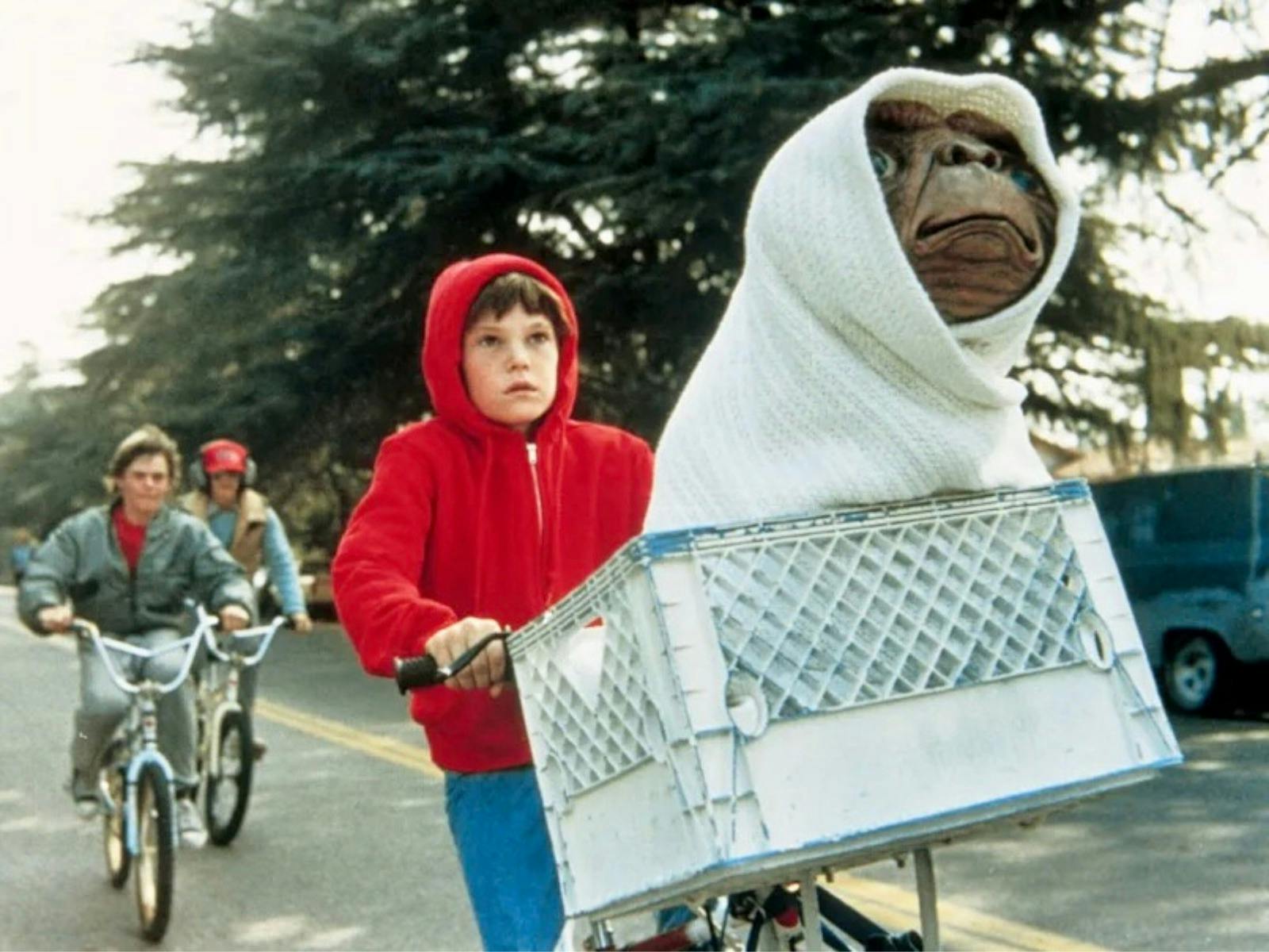 Three children on bicycles riding down a street lined by trees, with a hooded alien in the basket