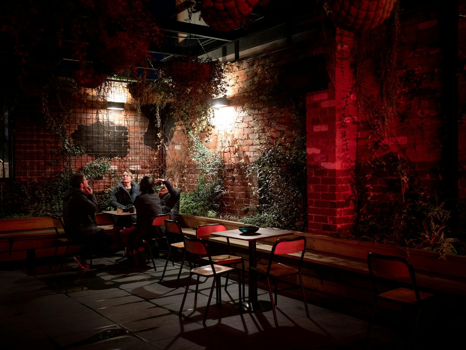 People seated at tables in an enclosed beer garden courtyard smile and laugh under red lighting