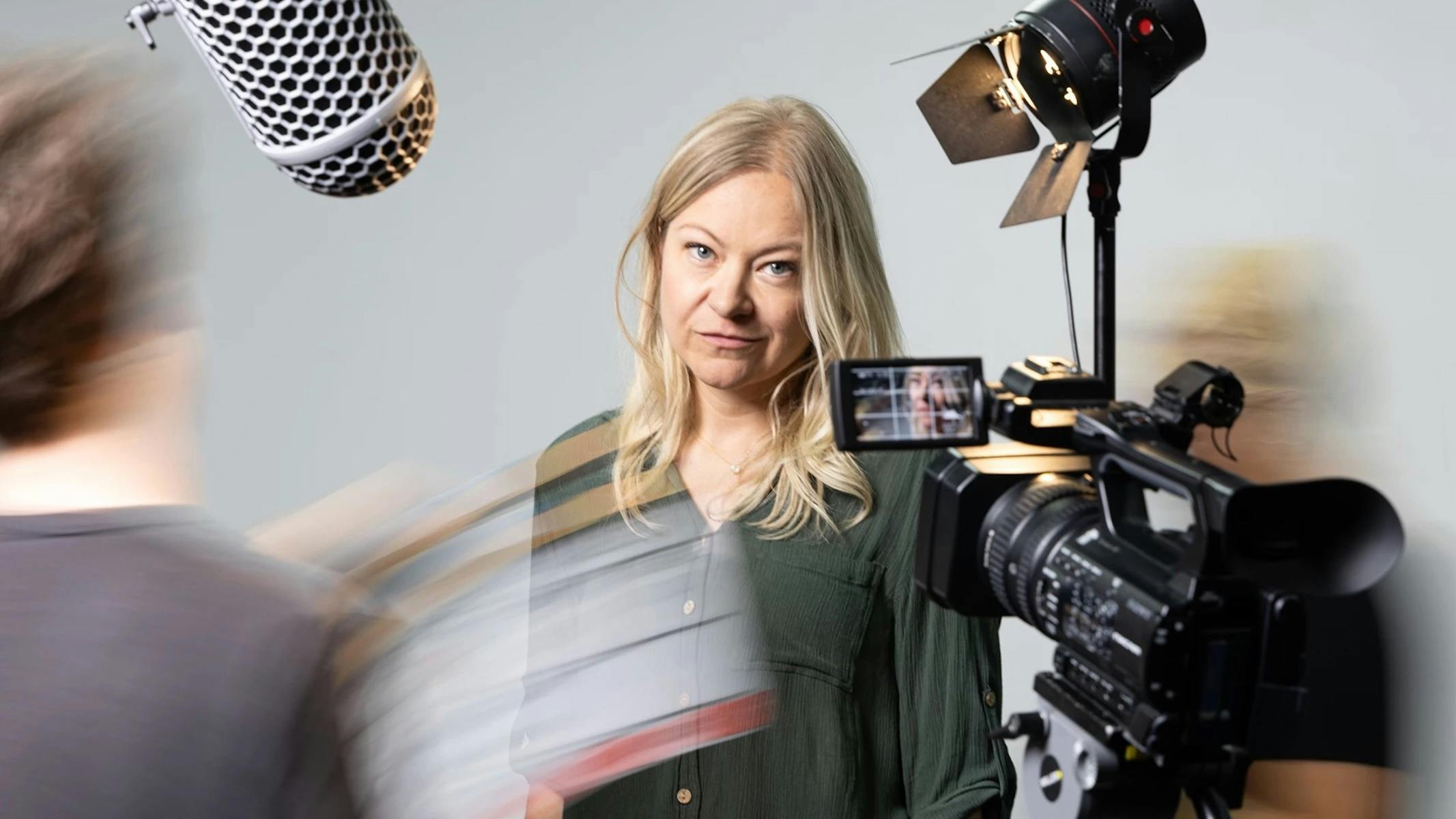 A woman with long blonde hair looks seriously at the camera, surrounded by filming equipment