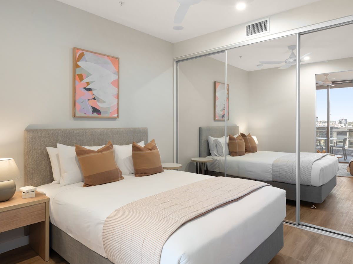 Queen-sized bed with cream coloured bedding and orange cushions, next to full length mirrored closet