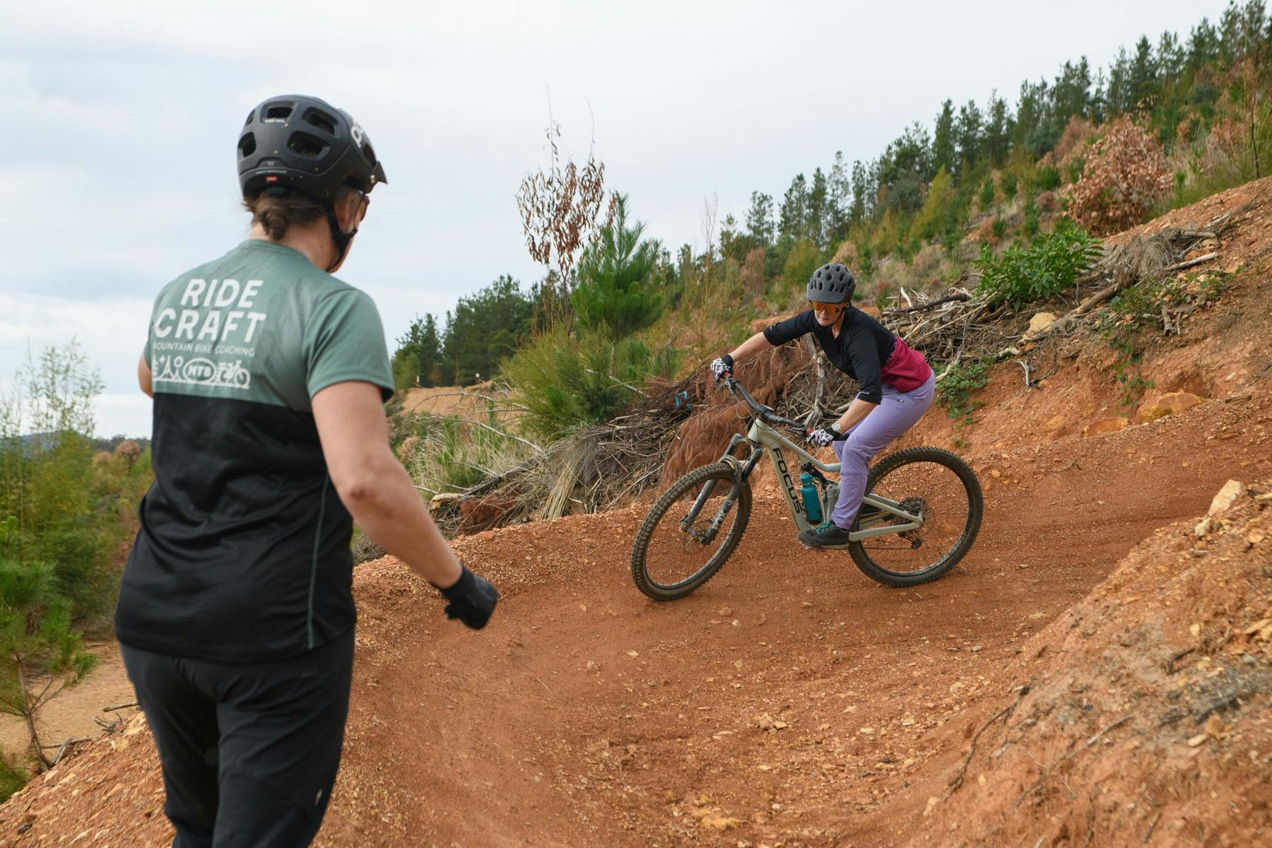 A MTB rider rides a corner while a coach looks on with her back to the camera