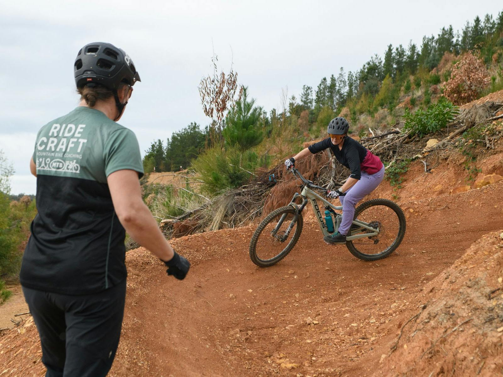 A MTB rider rides a corner while a coach looks on with her back to the camera