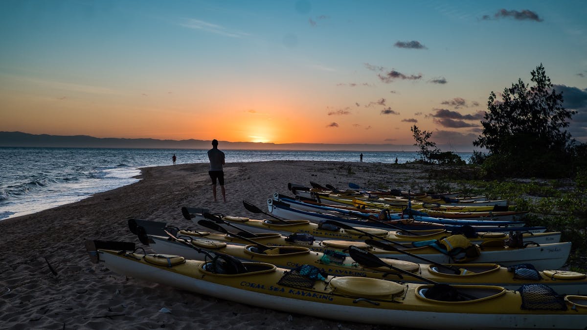 A person stands on Wheeler Island beach watching the sun set. The fleet of coral sea kayaks in fore