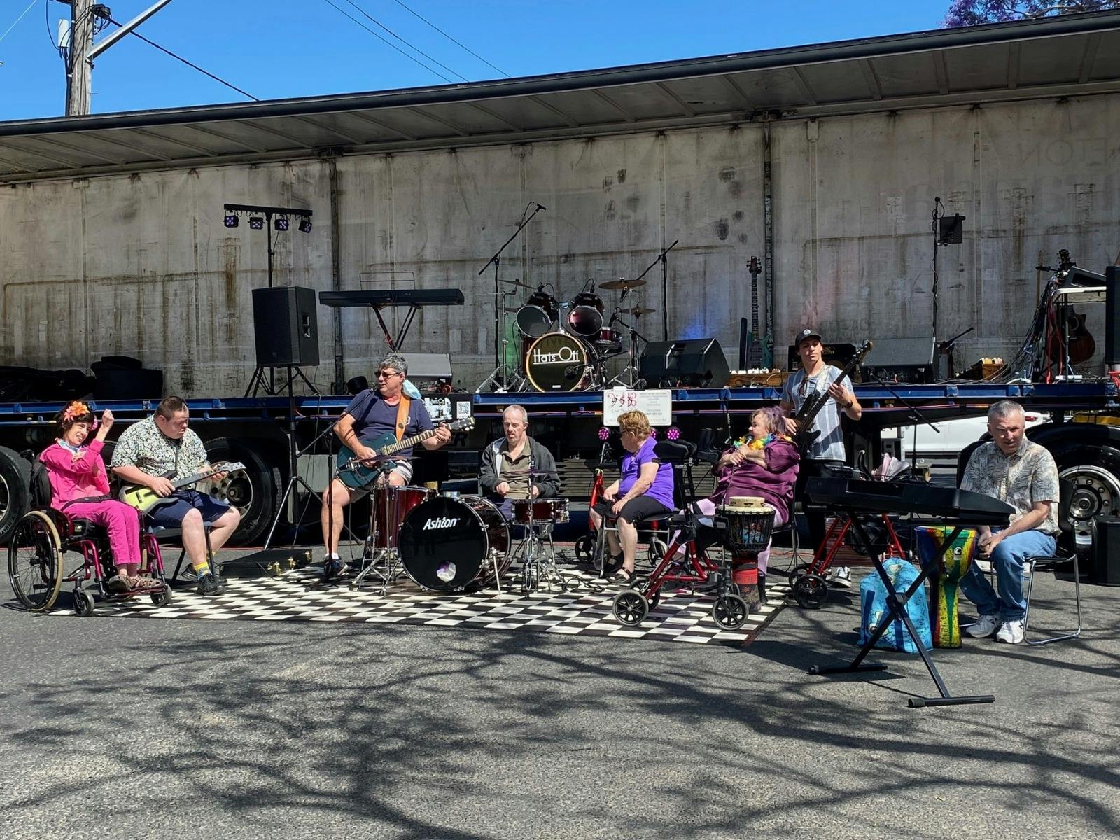 Community musicians perform outdoors in front of a stage mounted on a truck.