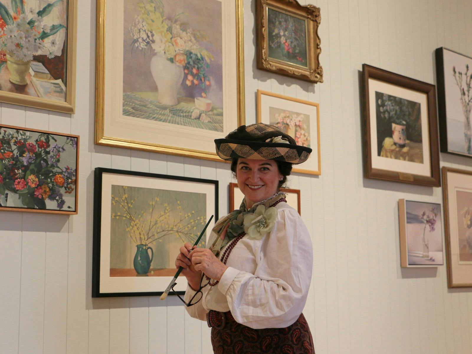 A woman dressed in old timey clothing stands in an exhibition space with paintings on walls