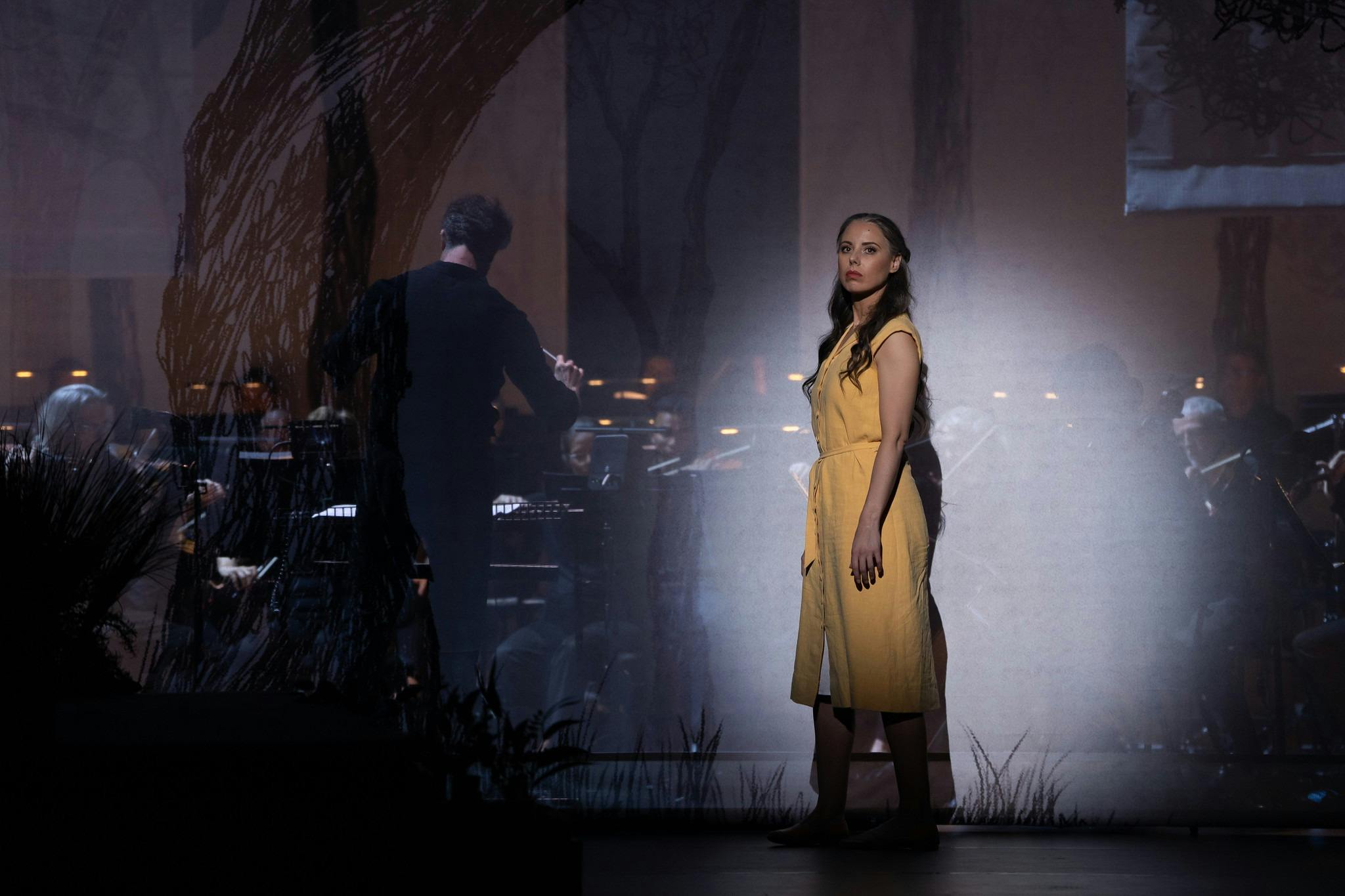 Performer in a yellow dress stands in spotlight beside an orchestra on stage.