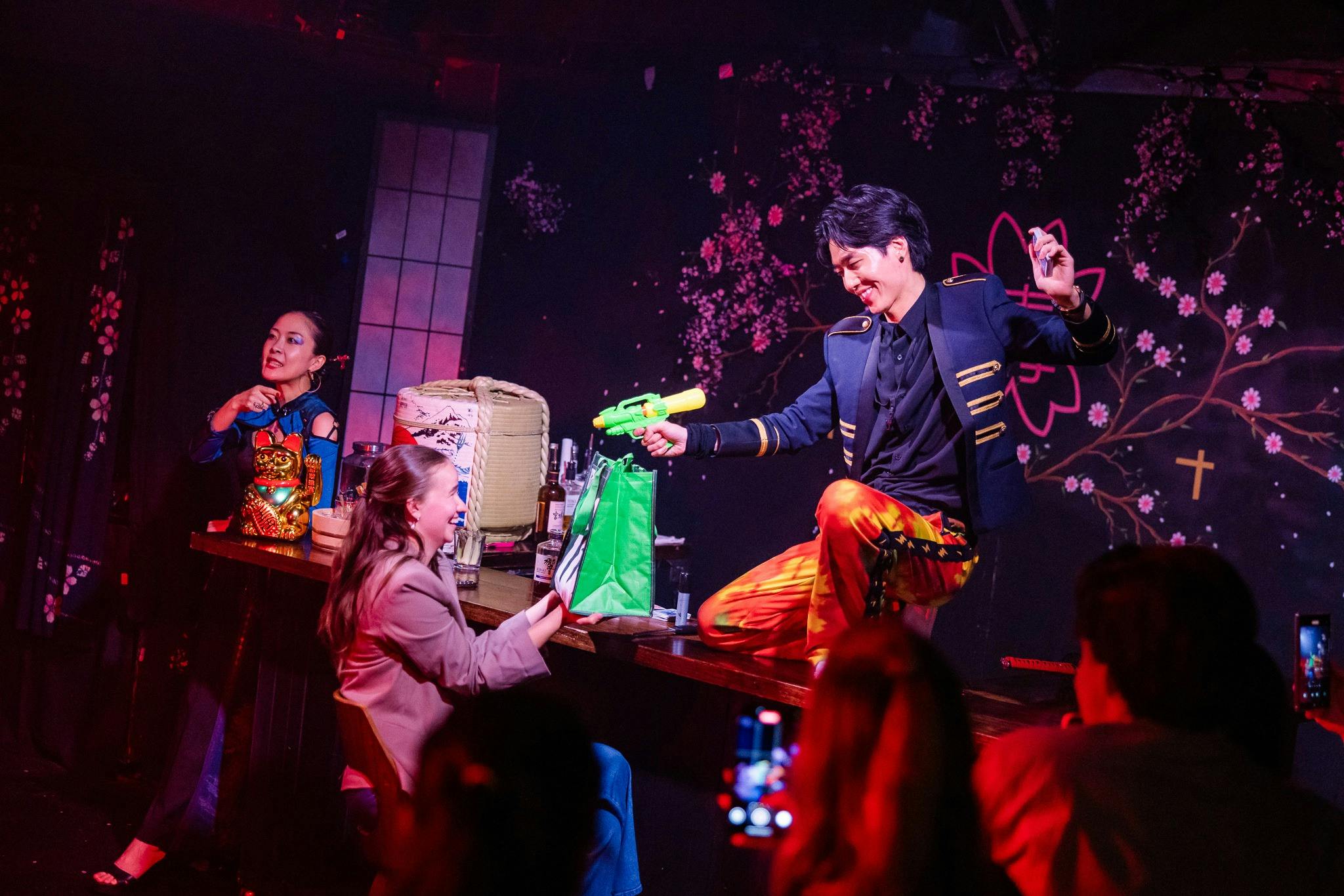 A magician points a colourful water gun at a laughing audience member
