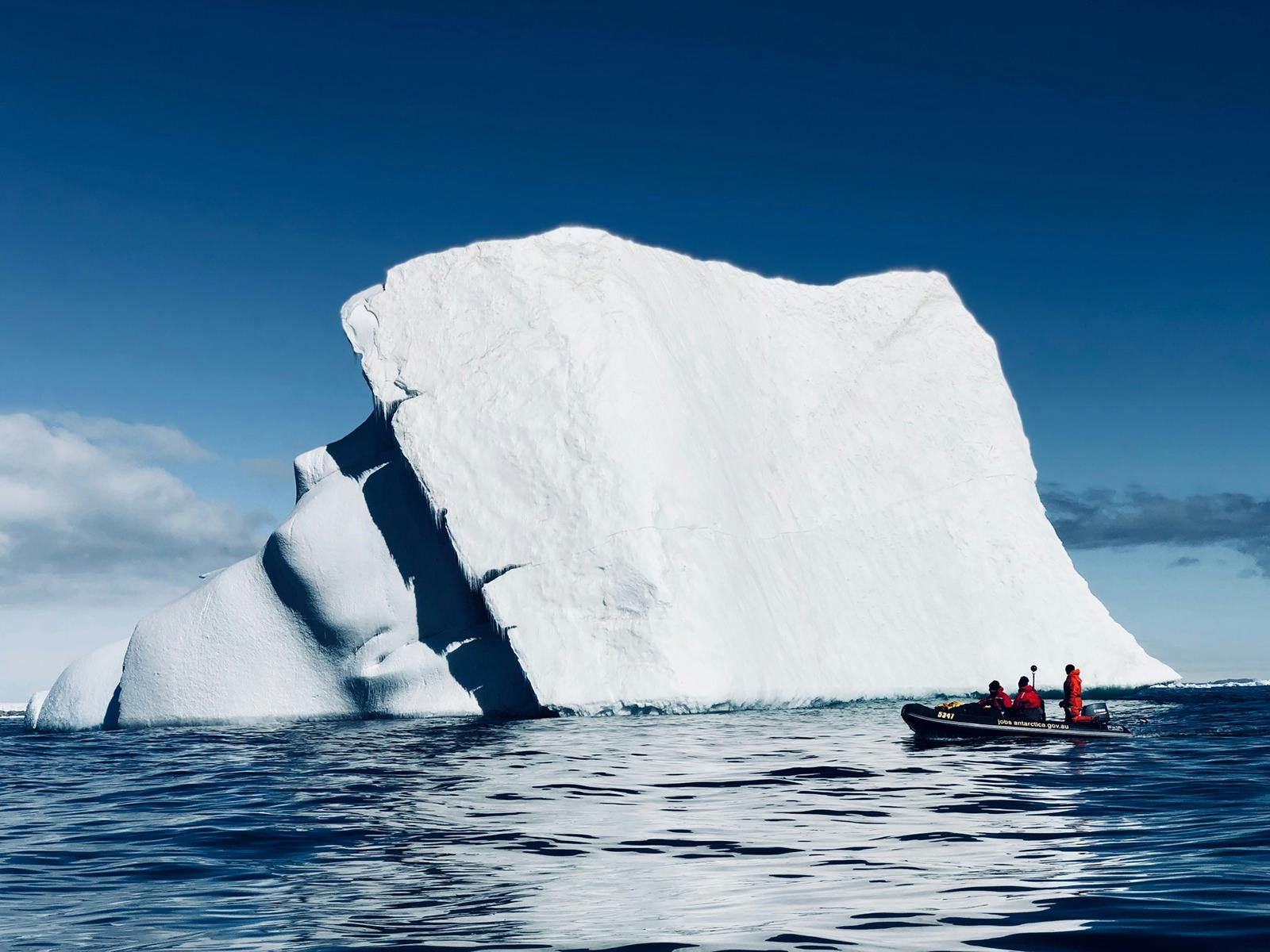 A zodiac boat with Antarctic expeditioners floats beside a large iceberg.
