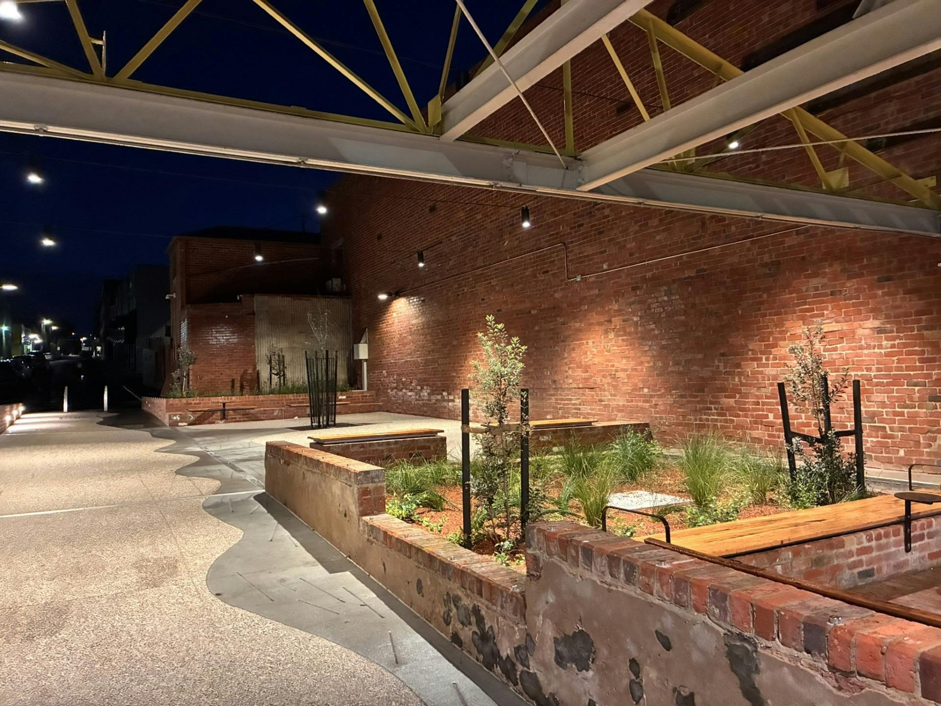 Illuminated brick laneway with landscaped garden beds, timber seats and exposed roof trusses.