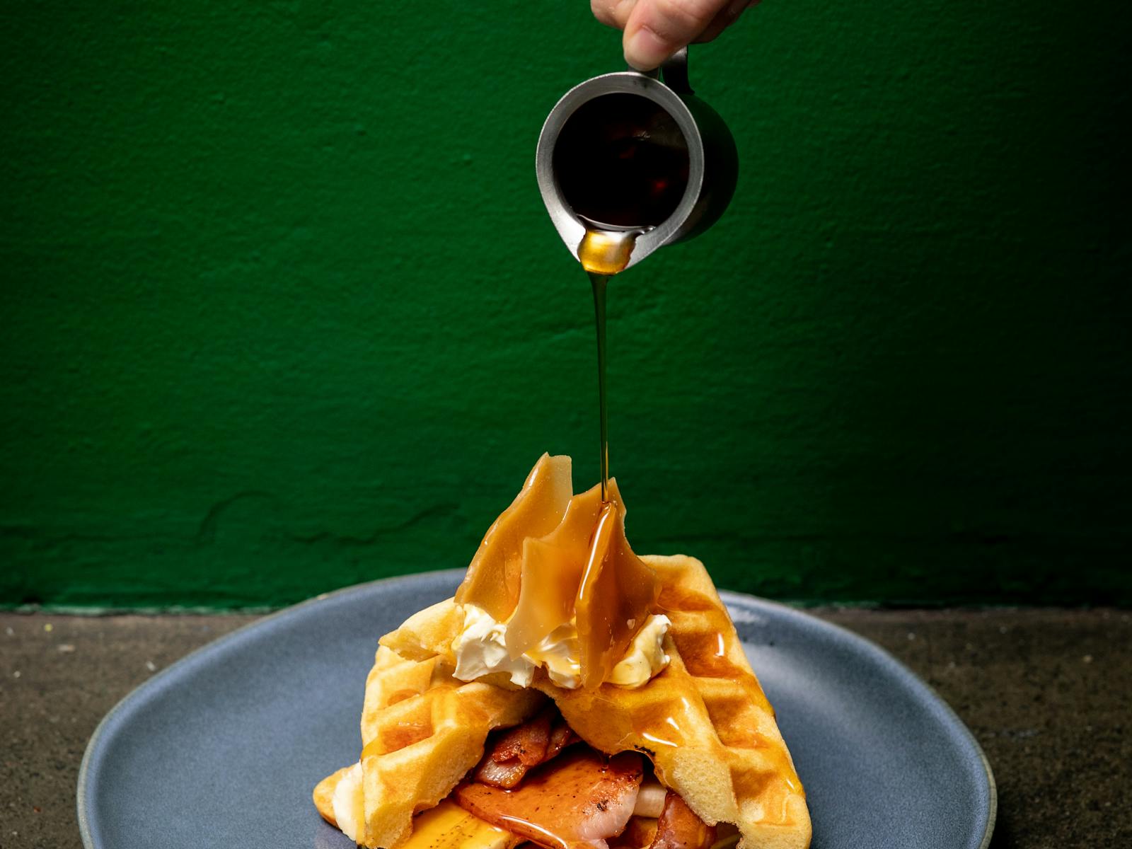 Honey waffles + bacon on a plate w cream. Jug above during maple syrup onto waffles