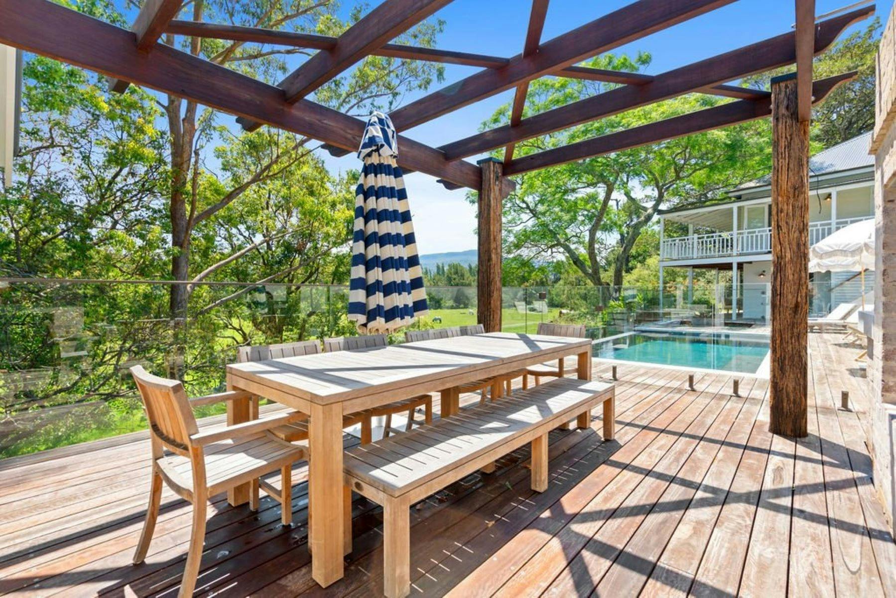 Image of outdoor dining table and chairs on deck. With pool and house in the background.