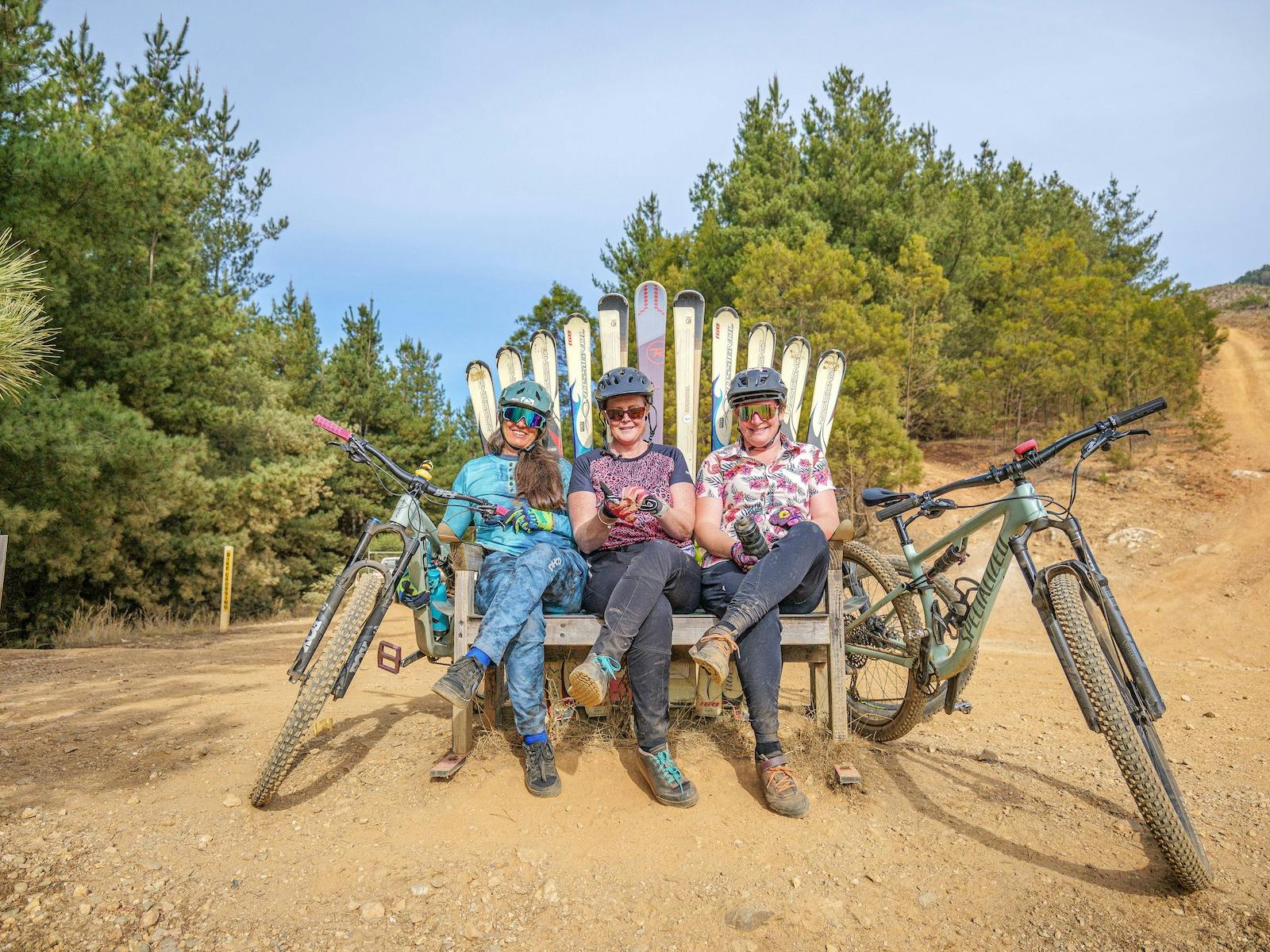 Three women in bike gear sit side by side on a large chair made of skis, with their bikes nearby.