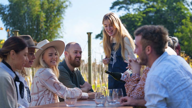 A group of people sitting outside at a winery sampling wine and enjoying each others company