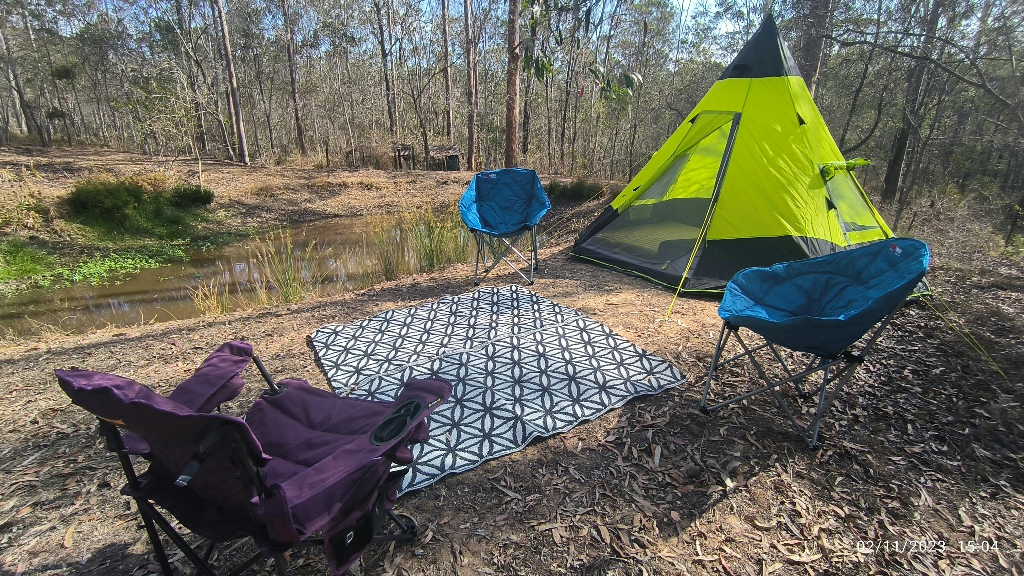 Tipi Tent setup in a bush setting with camping chairs and a mat overlooking a dam