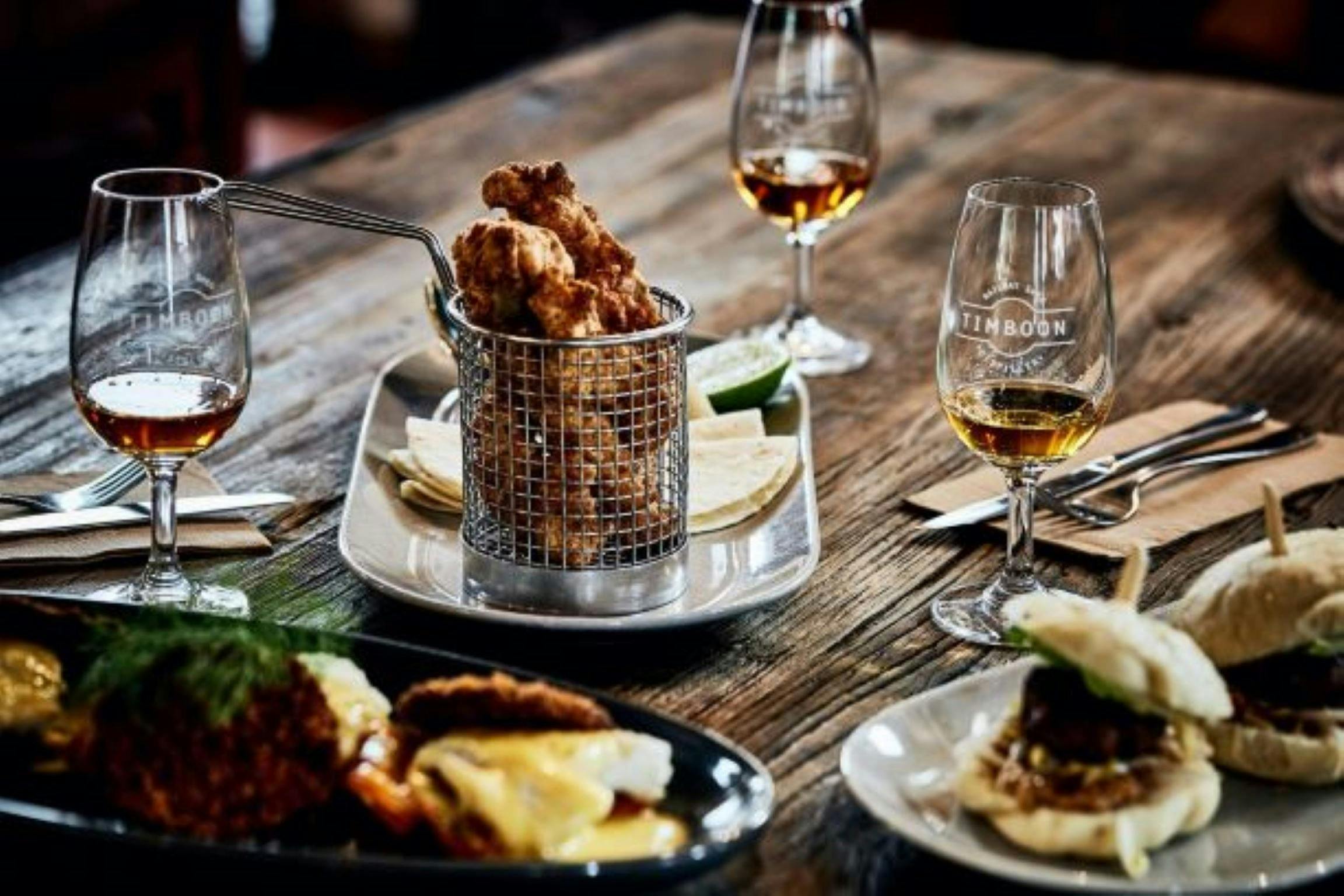 Fried chicken sits in a basket, four glasses of whisk surround it. Bao buns in the bottom right.