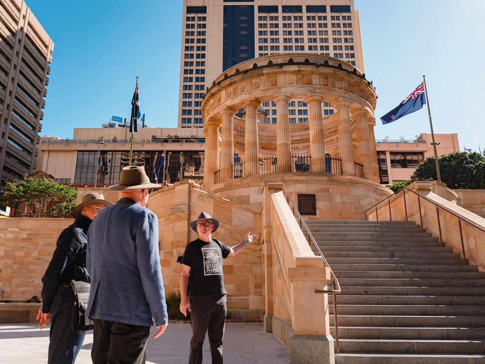 Two people stand in front of a Museum tour guide outside a large sandstone memorial with columns.
