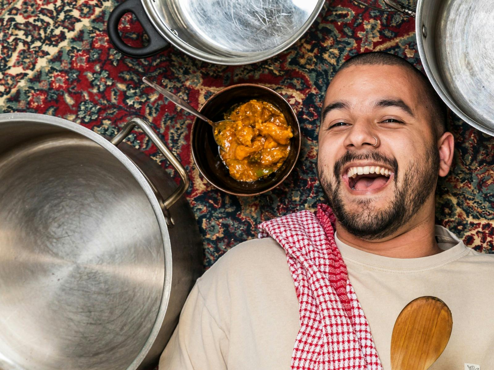 A person with a cheerful expression lies on a patterned rug next to a pot and a bowl of food.